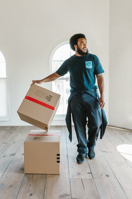 A man with dark curly hair and a beard is inside a bright, airy room with white walls and large arched windows, during a home relocation or furniture transport process. He is wearing a navy blue T-shirt with a patterned pocket and dark trousers, and is holding a cardboard box wrapped with a red packing strap. Two additional cardboard boxes are on the wooden floor next to him, one of which he is lifting or placing. The boxes are made of brown corrugated cardboard, some with visible shipping labels. The room appears to be part of a residential property, with natural light illuminating the scene. This image depicts a professional moves' loading or packing activity, representative of services offered by Man with Van Pimlico, as part of their removals and home relocation operations.