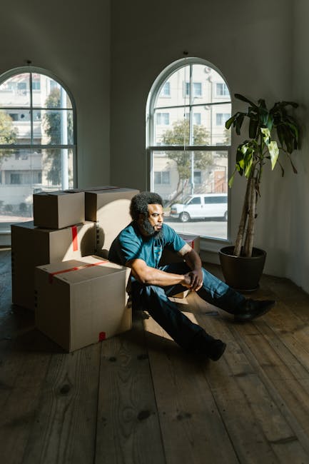A man with curly hair and medium skin tone sitting on a wooden floor inside a room with natural light coming through large arched windows shows him resting among several cardboard boxes, some of which are taped closed with red packing tape. The boxes are stacked around him, indicating a home relocation or moving process. He is wearing a blue T-shirt and dark pants, with his arms resting on his knees and a contemplative expression on his face. Behind him, a tall potted plant with large green leaves is positioned near the window, and outside the window, a street with parked cars and modern buildings is visible. This scene suggests a professional house removal or packing service, such as those offered by Man with Van Pimlico, during a furniture transport or packing stage of a move.