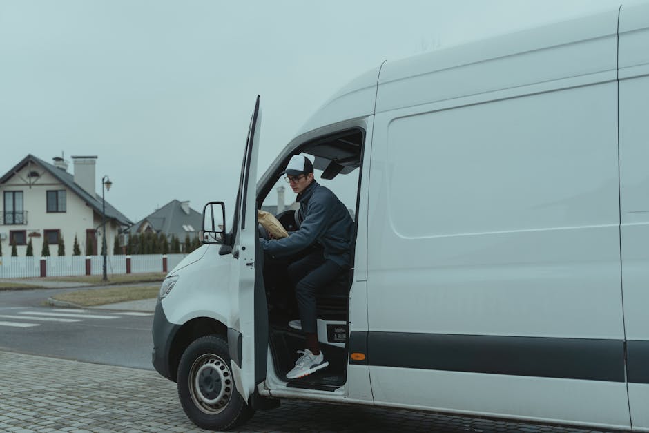 A man wearing a black cap, glasses, a dark jacket, and black trousers is sitting in the driver's seat of a white van, with the door open. He is holding a cardboard box with packing materials, preparing for a home relocation. The van is parked on a paved driveway near a residential street with houses featuring pitched roofs and chimneys. The background shows a cloudy sky, a sidewalk, and a streetlamp, indicating daytime. Inside the van, visible equipment includes a floor mat and straps for securing belongings during transportation. Man with Van Pimlico uses this vehicle for packing and moving furniture and boxes, supporting house removals and relocation services in the Pimlico area, with a focus on efficient furniture transport and loading processes.