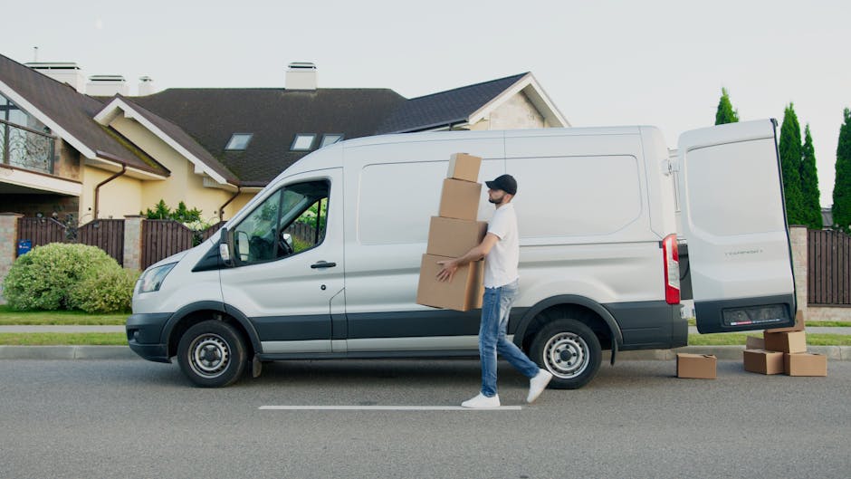 A man wearing a white t-shirt, jeans, and a black cap is loading three large cardboard boxes into the back of a white commercial van with sliding doors, parked on a residential street in Pimlico. The van belongs to Man with Van Pimlico, a house removal service, and is positioned on the roadway in front of a row of modern houses with pitched roofs, dormer windows, and a combination of brick and stucco exteriors. The open door reveals the interior of the van where the items are being packed carefully, and several additional cardboard boxes are placed on the pavement nearby. The scene captures the loading process during daylight hours, with greenery including bushes and tall trees visible behind the houses, emphasizing an organized home relocation or furniture transport operation involving packing materials, loading equipment, and a vehicle ready for moving services.