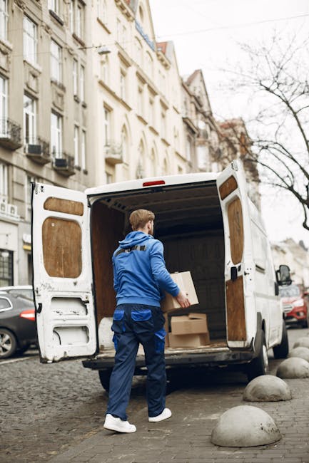 A man wearing a black cap, glasses, a dark jacket, and black trousers is sitting in the driver's seat of a white van, with the door open. He is holding a cardboard box with packing materials, preparing for a home relocation. The van is parked on a paved driveway near a residential street with houses featuring pitched roofs and chimneys. The background shows a cloudy sky, a sidewalk, and a streetlamp, indicating daytime. Inside the van, visible equipment includes a floor mat and straps for securing belongings during transportation. Man with Van Pimlico uses this vehicle for packing and moving furniture and boxes, supporting house removals and relocation services in the Pimlico area, with a focus on efficient furniture transport and loading processes.