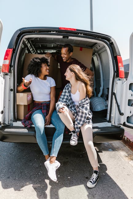 Two young women sit inside the open rear of a moving van during a home relocation, with one wearing a white T-shirt and jeans, and the other in a white top with a checked shirt tied around her waist. The woman on the left is gesturing with her hand while smiling, and the one on the right is looking at her with a smile. The van's cargo area contains cardboard boxes and packing materials, including a plastic-wrapped item and a rolled-up rug. The vehicle is parked outdoors on a sunny day, with the pavement and a curb visible. This scene illustrates the loading process during furniture transport and packing and moving activities, highlighting the involvement of [COMPANY_NAME] in professional removals services, supporting home relocation or house removals.