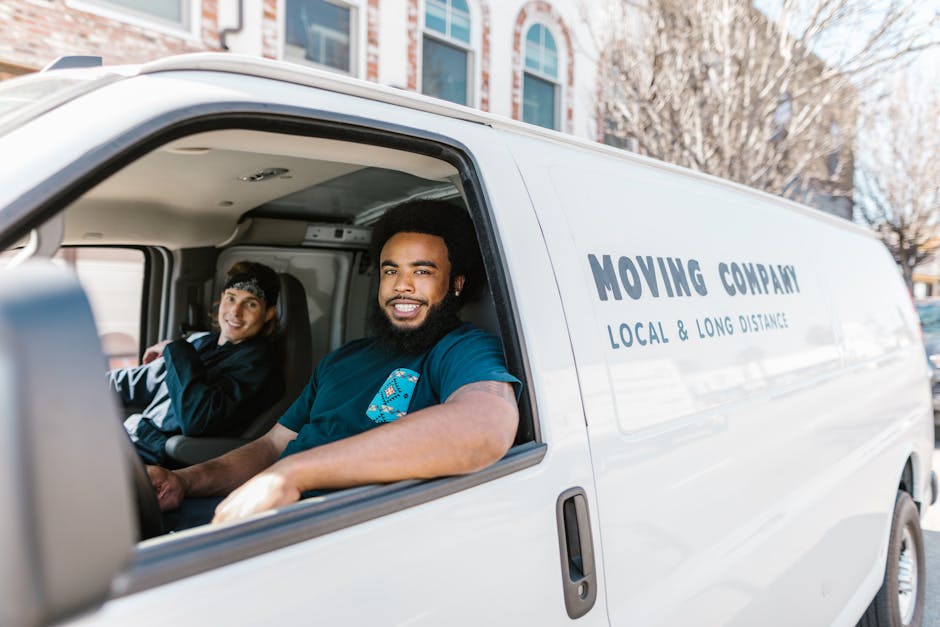 Inside the cabin of a white moving van belonging to Man with Van Pimlico, a male driver with a beard and dark hair is seated in the driver’s seat, smiling and looking towards the camera. Sitting beside him in the passenger seat is a woman with dark hair wearing a black jacket, also smiling. The van is parked outdoors on a city street in front of a multi-storey residential building with large windows and some trees with leafless branches. The side of the van displays the company name, 'MOVING COMPANY', along with the description 'LOCAL & LONG DISTANCE REMOVALS'. The image captures the loading process or travel phase of a house relocation, with natural daylight illuminating the scene. The vehicles and the individuals suggest they are engaged in packing, furniture transport, or moving logistics as part of professional residential or commercial move services offered by Man with Van Pimlico.