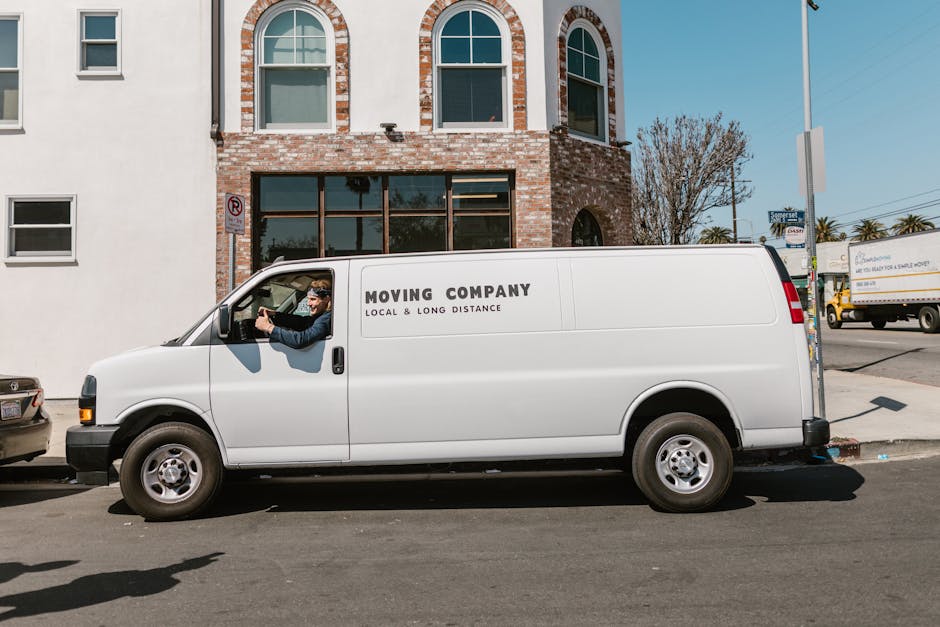 A white moving van operated by Man with Van Pimlico parked on the street outside a brick-built residential building with large windows and arched top windows on the upper floor. The van has the text 'Moving Company Local & Long Distance' printed on its side. The driver, wearing glasses and a dark jacket, is sitting in the cab, holding the steering wheel with both hands. In the background, there are other vehicles, including a yellow truck and a car, along with bare trees and a clear blue sky. The scene captures the loading process or home relocation logistics, with the van positioned adjacent to the building for easy access, and the overall environment suggests a professional furniture transport or house move conducted by Man with Van Pimlico.
