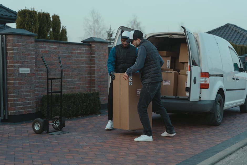 A man with a beard, dressed in a blue uniform, is standing outside a white removal van on a cobblestone street in Pimlico, during a home relocation process. He is holding a cardboard box, preparing to load it into the van, which has its rear doors open to reveal an interior space lined with wooden panels for furniture transport. On the ground near him are additional cardboard boxes, some placed on the pavement and others positioned close to the van's open door. The scene is set against a backdrop of multi-storey residential buildings with traditional architecture, featuring white facades, black iron balconies, and large windows. Parking cars are visible along the street, and leafless trees indicate a colder season. The lighting suggests daytime with natural daylight illuminating the scene, capturing the packing and moving process overseen by Man with Van Pimlico as part of their professional removals services.