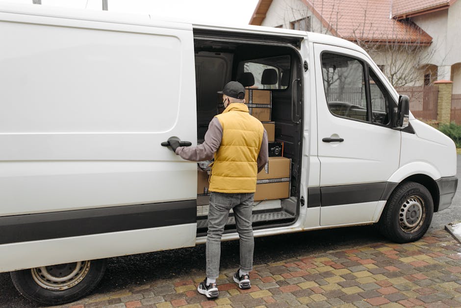 A man wearing a yellow padded vest, grey sweatshirt, grey jeans, and a black cap is standing outside in front of a white cargo van with sliding door open, preparing to load or unload packed cardboard boxes of various sizes that are stacked inside the van near the open doorway. The van is parked on a paved driveway adjacent to a residential area with brick paving, and there are houses with pitched roofs, wooden fencing, and leafless trees visible in the background. The scene captures the logistics of home relocation, with the man from Man with Van Pimlico involved in a furniture transport or packing and moving process, utilizing equipment such as the van to facilitate house removals or studio moves.