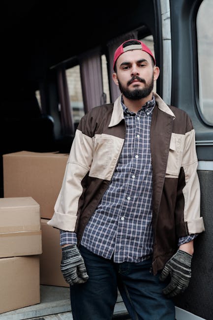 A young man with a beard and wearing a red baseball cap, a plaid shirt, and a beige and brown work jacket stands inside a van used for house removals, with his hands in his pockets and a neutral expression. Behind him are stacked cardboard boxes ready for transportation, some wrapped in clear plastic film for protection. The van interior features dark panels and a floor suitable for secure loading and unloading of furniture and packing materials. Natural lighting illuminates the scene, highlighting the furniture and packaging as part of the home relocation process undertaken by Man with Van Pimlico, specializing in removals for flats and studios, with emphasis on loading and transport logistics.