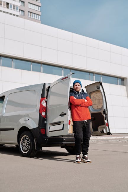 A man wearing a yellow padded vest, grey sweatshirt, grey jeans, and a black cap is standing outside in front of a white cargo van with sliding door open, preparing to load or unload packed cardboard boxes of various sizes that are stacked inside the van near the open doorway. The van is parked on a paved driveway adjacent to a residential area with brick paving, and there are houses with pitched roofs, wooden fencing, and leafless trees visible in the background. The scene captures the logistics of home relocation, with the man from Man with Van Pimlico involved in a furniture transport or packing and moving process, utilizing equipment such as the van to facilitate house removals or studio moves.