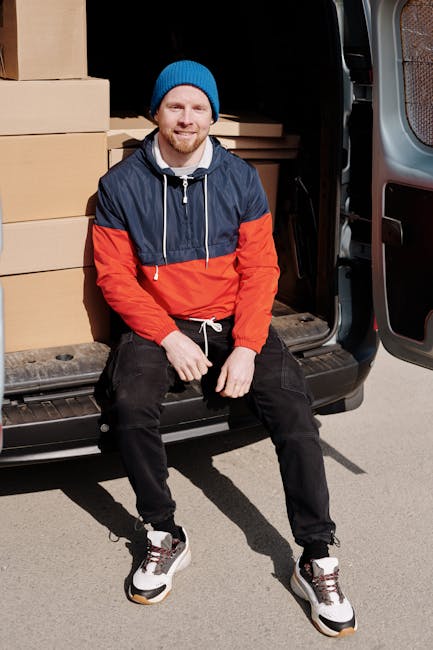 A male removal driver with a beard and wearing a red cap, brown and beige work jacket, and gloves, standing inside a van with an open side door. Behind him are several cardboard moving boxes, some sta