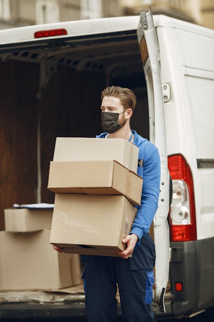 A man wearing a black face mask and a blue jacket is standing at the rear of a white moving van, holding three cardboard boxes of varying sizes, with one box stacked on top of another. The van's rear doors are open, revealing a partially visible interior filled with more cardboard boxes and packing materials arranged for a home relocation. The man appears to be in the process of loading or unloading the boxes, which are wrapped in clear plastic for protection during transit. Outside the van, on the pavement, additional cardboard boxes and packing supplies are visible, indicating an active packing and moving process. The environment suggests an urban area, with nearby buildings visible in the background, providing context for local house removals handled by Man with Van Pimlico. This scene illustrates professional furniture transport and packing efforts during a same-day last-minute move, emphasizing the logistics involved in home relocation services.
