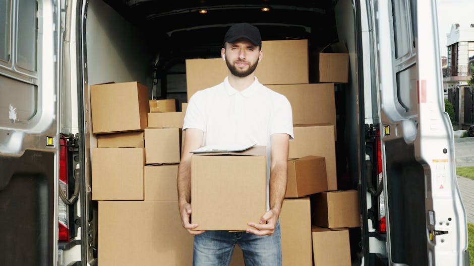 A man wearing a black cap, white polo shirt, and blue jeans stands inside the open rear of a white van, holding a cardboard box with both hands. The van is packed with numerous cardboard boxes of various sizes, some stacked on top of each other, filling the cargo area. The boxes are sealed with brown packing tape and lined up neatly, suggesting an organized packing and loading process typical of house removals. The van is parked outdoors near a residential area, with visible pavement and nearby buildings in the background. The lighting indicates daytime, with natural sunlight illuminating the scene. This image depicts furniture transport and packing during a home relocation, as conducted by Man with Van Pimlico, a specialist in last-minute same-day removals and professional moving services.