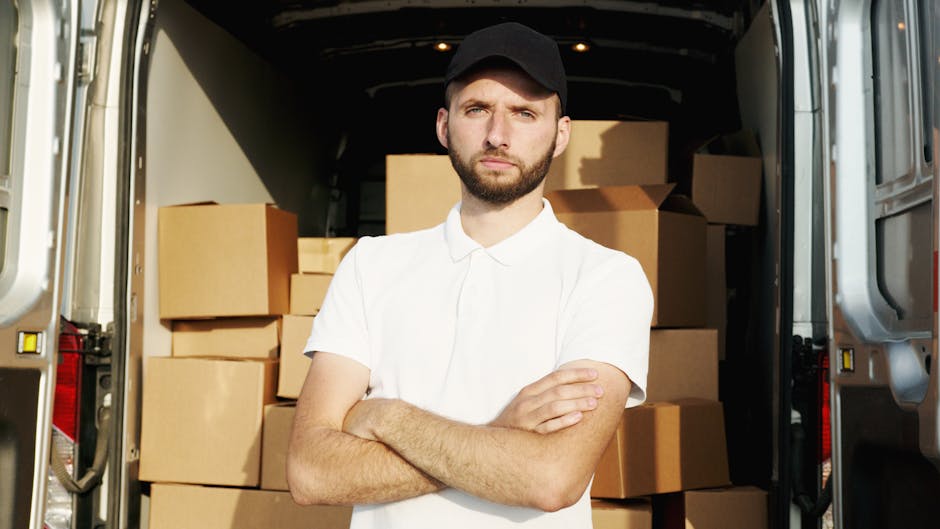A man wearing a black face mask and a blue jacket is standing at the rear of a white moving van, holding three cardboard boxes of varying sizes, with one box stacked on top of another. The van's rear doors are open, revealing a partially visible interior filled with more cardboard boxes and packing materials arranged for a home relocation. The man appears to be in the process of loading or unloading the boxes, which are wrapped in clear plastic for protection during transit. Outside the van, on the pavement, additional cardboard boxes and packing supplies are visible, indicating an active packing and moving process. The environment suggests an urban area, with nearby buildings visible in the background, providing context for local house removals handled by Man with Van Pimlico. This scene illustrates professional furniture transport and packing efforts during a same-day last-minute move, emphasizing the logistics involved in home relocation services.