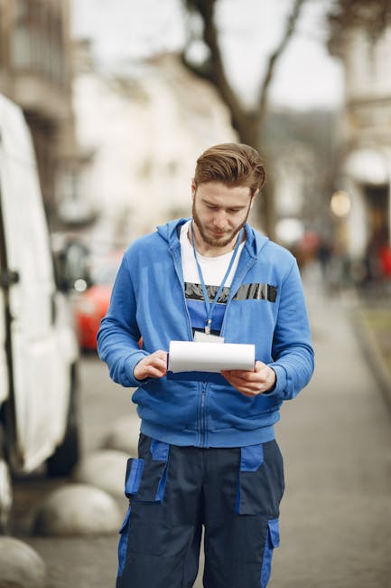 A young male relocation specialist from Man with Van Pimlico stands outdoors on a city street during daytime, reading a clipboard with notes. He is dressed in a blue work hoodie with black and grey ac