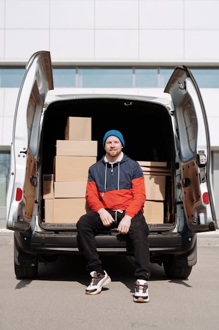 A man with a beard, dressed in a red and navy blue jacket, black trousers, white sneakers, and a blue beanie sits on the edge of an open van trunk, which is loaded with multiple brown cardboard boxes of varying sizes, some stacked on top of each other. The boxes are wrapped in clear plastic wrap to secure them for transport during a home relocation or moving process. The van is parked on a paved surface in front of a modern building with a white tiled wall and large glass windows, indicating an urban environment. Behind the man, inside the vehicle, additional packaging materials such as bubble wrap and packing paper can be seen, supporting the packing and moving activities carried out by [COMPANY_NAME], specializing in removals. The lighting is natural, suggesting daytime, and the scene captures the loading process involved in furniture transport and house removals, with the man actively involved in the logistics of a residential move in Pimlico.