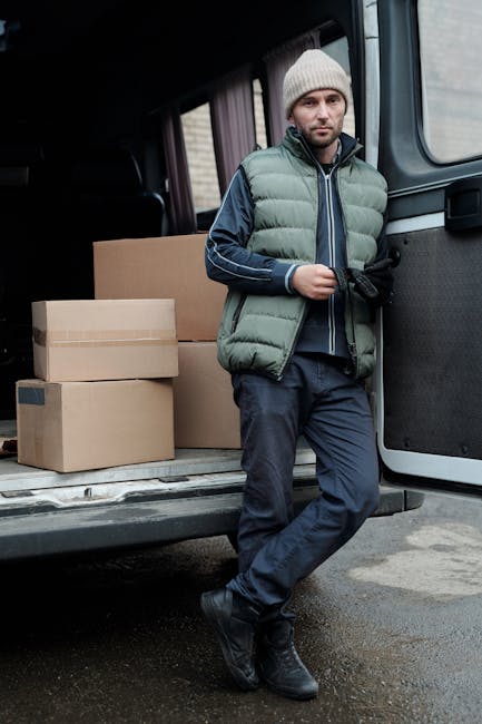 A man wearing a grey knit beanie, a green puffer vest over a navy hoodie, and dark trousers stands outside a black van, which is parked on a paved area near a residential building. The back doors of the van are open, revealing several stacked cardboard boxes of various sizes, some secured with plastic wrap, ready for home relocation. The man is holding a black item in one hand and leans against the side of the van with his other arm. The scene depicts a loading process during furniture transport or packing and moving activities, typical of professional removals services. The environment appears overcast, with natural daylight illuminating the scene, indicating an outdoor moving operation. This image exemplifies the logistics involved in house removals, with items packed in boxes prepared for transportation, aligned with moving companies like Man with Van Pimlico.