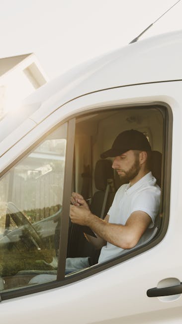 A man with a beard, dressed in a red and navy blue jacket, black trousers, white sneakers, and a blue beanie sits on the edge of an open van trunk, which is loaded with multiple brown cardboard boxes of varying sizes, some stacked on top of each other. The boxes are wrapped in clear plastic wrap to secure them for transport during a home relocation or moving process. The van is parked on a paved surface in front of a modern building with a white tiled wall and large glass windows, indicating an urban environment. Behind the man, inside the vehicle, additional packaging materials such as bubble wrap and packing paper can be seen, supporting the packing and moving activities carried out by [COMPANY_NAME], specializing in removals. The lighting is natural, suggesting daytime, and the scene captures the loading process involved in furniture transport and house removals, with the man actively involved in the logistics of a residential move in Pimlico.