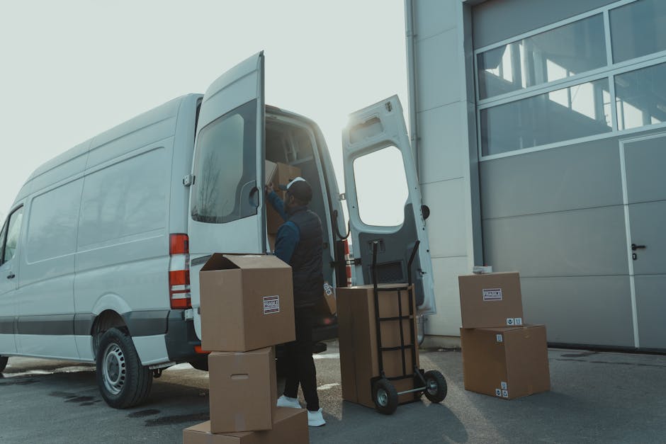 A professional mover from Man with Van Pimlico is shown loading cardboard boxes and packing materials into the rear of a white cargo van outside a modern building. The mover is wearing a dark vest and cap, handling a box that is being lifted onto the vehicle's open rear door. Several other boxes, some sealed with packaging labels, are positioned on the pavement nearby, along with a small hand trolley and a lifting strap for securing load stability. The scene takes place during daylight hours, with natural light illuminating the urban environment, highlighting the process of home relocation involving furniture transport and packing. The van is parked close to a grey industrial-style building with large windows, indicating an urban residential or commercial setting suitable for house removals services.