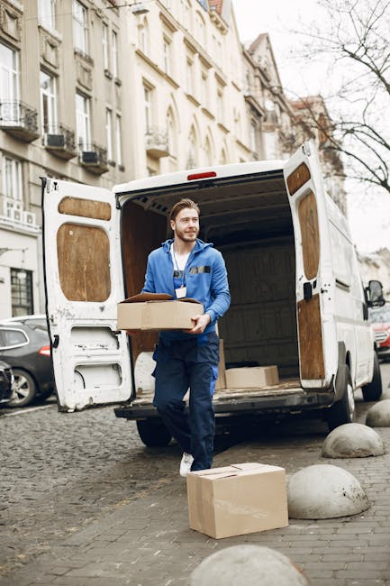 A professional mover from Man with Van Pimlico is shown loading cardboard boxes and packing materials into the rear of a white cargo van outside a modern building. The mover is wearing a dark vest and cap, handling a box that is being lifted onto the vehicle's open rear door. Several other boxes, some sealed with packaging labels, are positioned on the pavement nearby, along with a small hand trolley and a lifting strap for securing load stability. The scene takes place during daylight hours, with natural light illuminating the urban environment, highlighting the process of home relocation involving furniture transport and packing. The van is parked close to a grey industrial-style building with large windows, indicating an urban residential or commercial setting suitable for house removals services.